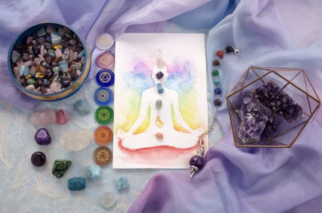 A vibrant drawing of a person in meditation, outlined in rainbow watercolors representing the aura. On the left side, there is a bowl filled with an assortment of colorful crystals, including seven flat stones etched with chakra symbols, each corresponding to the respective chakra's color. On the right side, two large amethyst crystals are displayed within a wire pyramid. This image represents the harmonizing and balancing qualities of Reiki therapy in connection with the chakras and energetic healing.